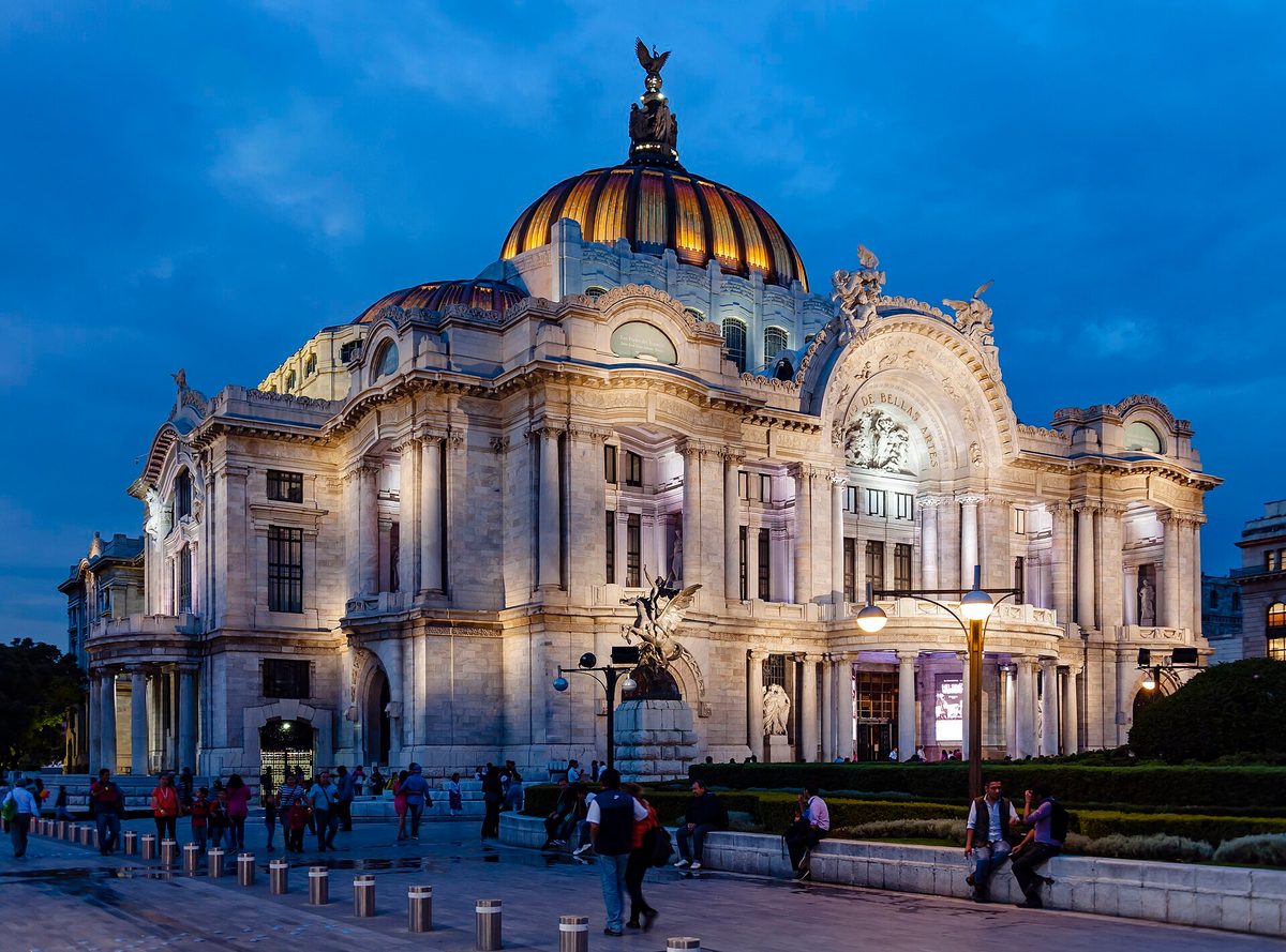 The Palacio de Bellas Artes in Mexico City at dusk, its domed marble facade lit up as people walk past